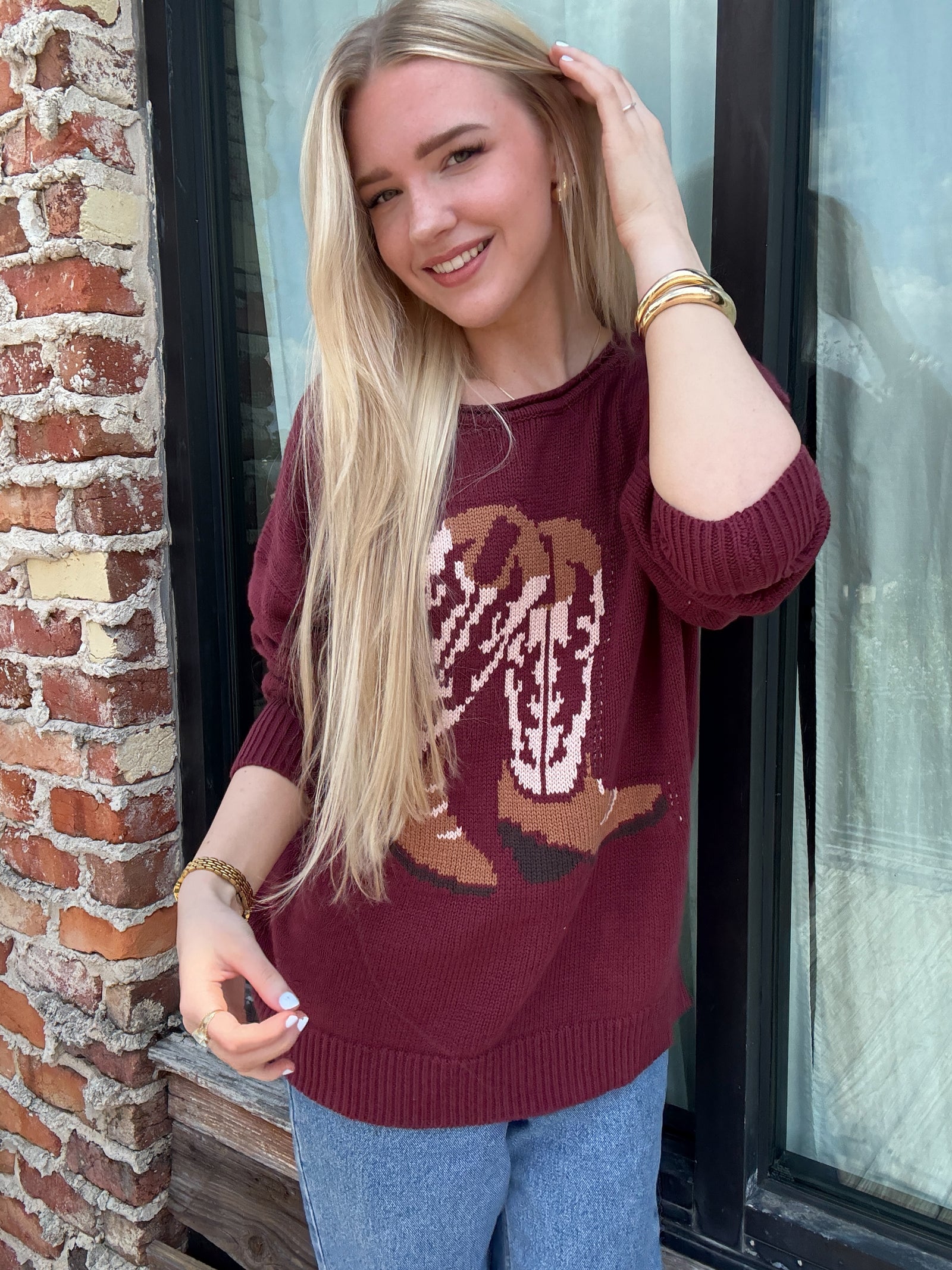 Woman wearing a maroon sweater with cowboy boot design against a brick wall outside of Addy & Ry Boutique in Spring Texas